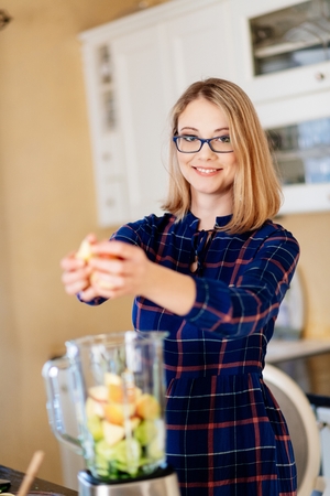 Young woman putting fruits and vegetables into the eletrical blender. Woman preparing delicious healthy smoothieの写真素材