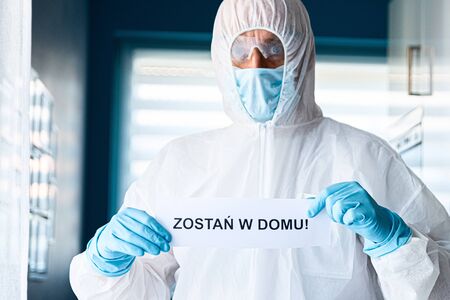 Man in protective anti virus suit and mask holding card with message stay at home in Polish languageの写真素材