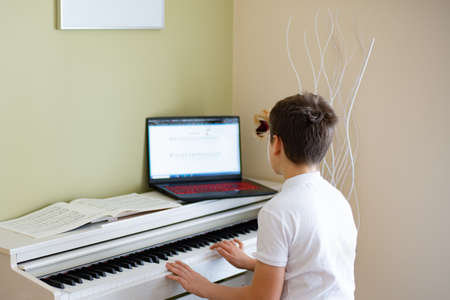 Young boy playing the white digital piano looking at the notes in the laptop.の写真素材