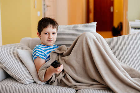 A young boy sits on the sofa under the blanket and switches the channels on television.の写真素材