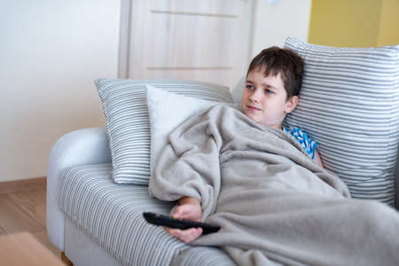 A young boy is lying on the sofa under a blanket, with the remote control in his hand, watching TV.の写真素材