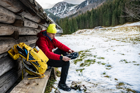 A man tourist in a yellow cap, red down jacket and yellow backpack puts crampons on his shoes.の写真素材
