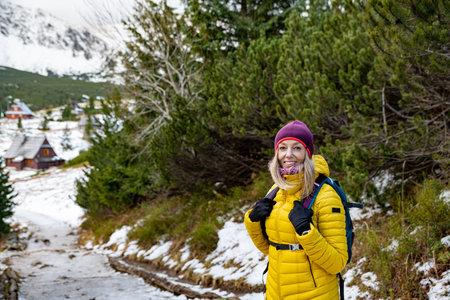A smiling woman hiker in a yellow down jacket with a backpack wanders the mountain trail in winter.の写真素材