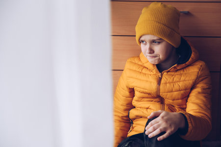 A sad teenager with a yellow jacket and cap sits by the window.の写真素材
