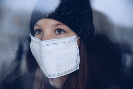 A pretty girl is standing by the window in a protective mask.の写真素材