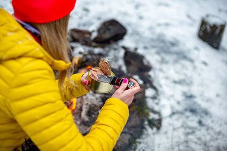 A young woman in a yellow jacket holds an open can of canned food by a campfire in a forest in the mountains.の写真素材