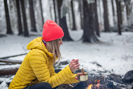 A blonde woman in a yellow jacket holds an open can of canned food in the woods by the fire.の写真素材