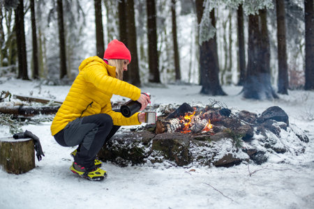 A blonde tourist in a yellow jacket pours hot tea into a metal mug by the fire.の写真素材