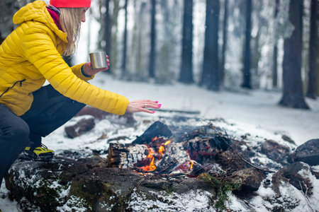 A blonde tourist in a yellow down jacket drinks tea and warms her hands by the fireの写真素材