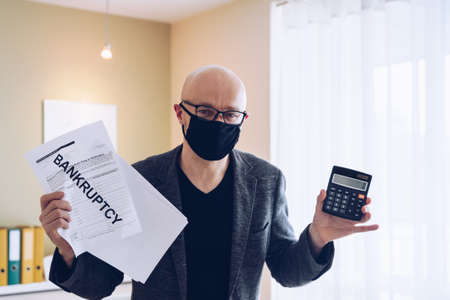 A man in a protective mask with a bankruptcy document and a calculator in his hand during a...