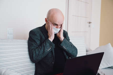 Businessman in protective mask sitting on the sofa he sees terrifying stock market results on the laptop.の写真素材