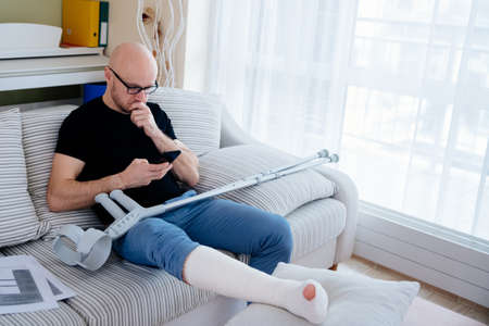 A man with glasses with a broken leg is sitting on the couch at home. Calls the insurer for compensation.の写真素材