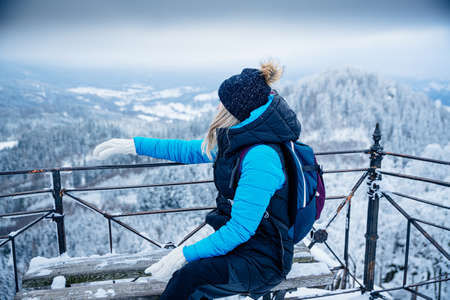 A blonde woman in a blue jacket sits on the observation deck and admires the snow-capped peak of the mountains.の写真素材