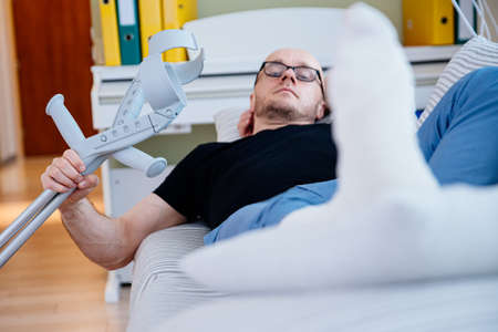 The man with the glasses is lying on the sofa at home with his leg in plaster.の写真素材