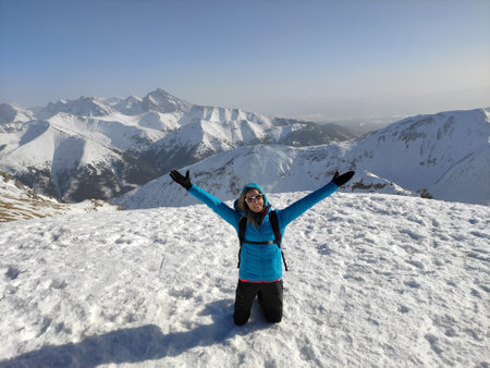 Happy female tourist in a blue jacket and arms raised up on top of a mountain.の写真素材