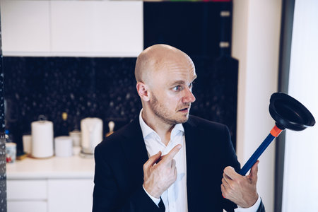 Handsome man in suit holding plunger in hand in kitchen. Drain problems.の写真素材