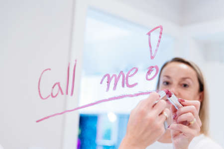 Blonde woman writes the words call me on the mirror with red lipstick.の写真素材