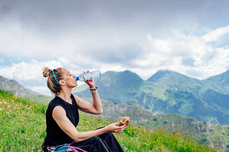 Hiker woman sitting, drinking water and eating sandwich, resting on the grass in the mountains.の写真素材