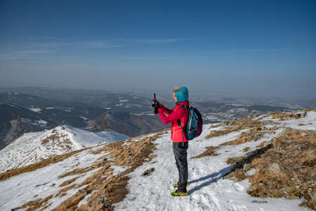 A tourist in a red jacket and backpack takes a photo with a phone on a beautiful mountain trail.の写真素材