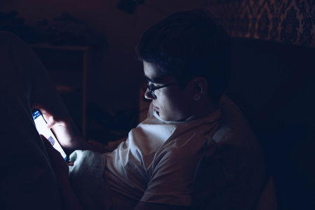 Teenager with glasses uses the phone in bed at night.の写真素材