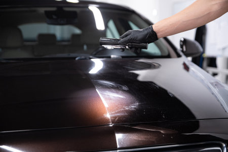 Employee of the car detailing studio conducts an inspection of the car paint after polishing the...