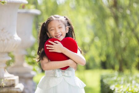 Little girl in white dress smiling and hug red heart pillow in garden, Valentine kid with heart in garden concept for valentine dayの写真素材