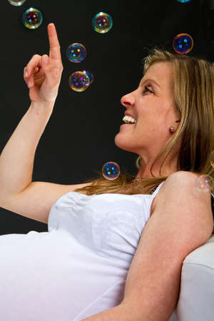 Young fresh pregnant woman with blow bubbles  sitting on a couch over black background.の写真素材
