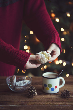 girl hand in red sweater holding holding baked cookie near by kitchen table with cookies and warm coffee drink. christmas breakfast table with bokeh lightsの写真素材