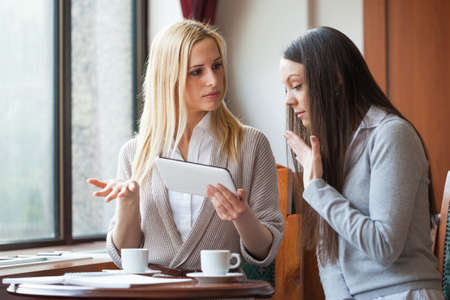 Two women drinking coffee and using digital tablet in cafeの写真素材