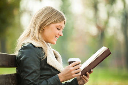 Young woman reading book and drinking coffee to go in parkの写真素材