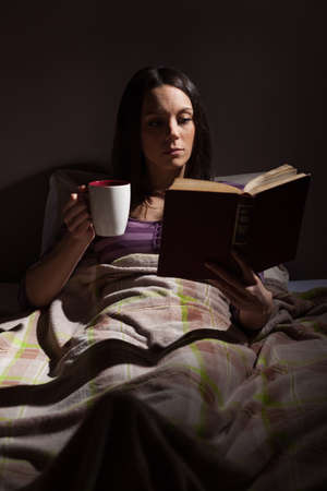 Young woman relaxing in her bed before sleep. She is reading a book and drinking tea.の写真素材