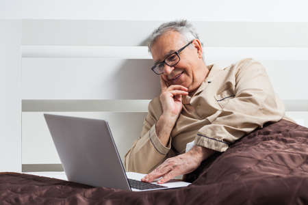 Happy senior man is lying in bed and using laptopの写真素材