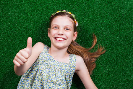 Happy little girl is lying on artificial grass and showing thumb up.の写真素材