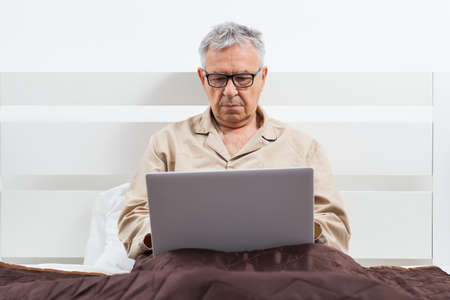 Senior man is lying in bed and using laptop.