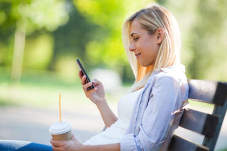 Young happy woman is sitting in park and using smartphone.の写真素材