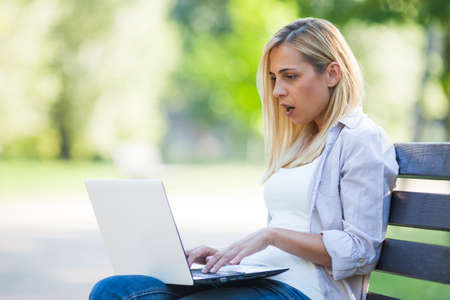 Young woman is sitting in park and reading news on laptop. She is surprised.の写真素材