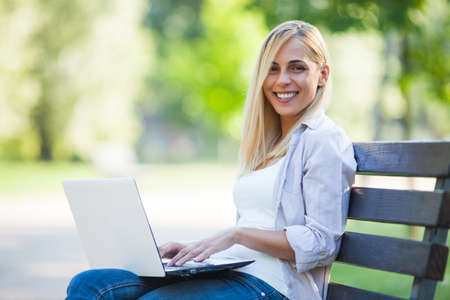 Young happy woman is sitting in park and using laptop.の写真素材
