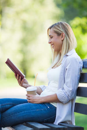 Young happy woman is sitting in park. She is reading book and drinking coffee to go.の写真素材
