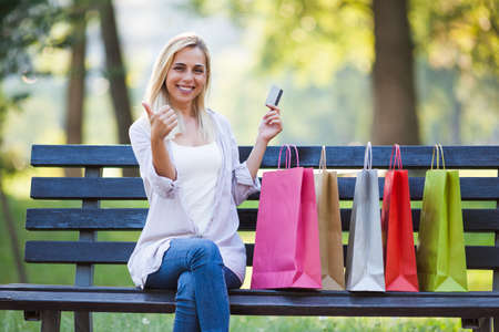 Young happy woman sitting in park after shopping and holding credit card.の写真素材