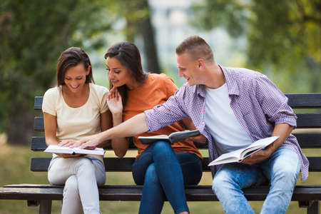 Three students are sitting on bench in park and learning.の写真素材
