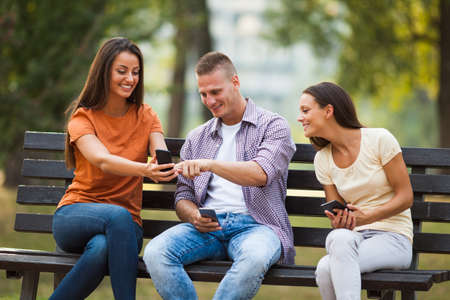 Three friends are sitting on bench in park and using smartphones.の写真素材