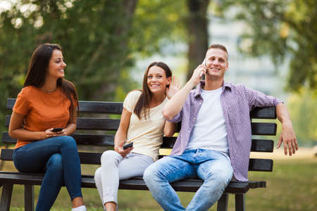 Three friends are sitting on bench in park and using smartphones.の写真素材