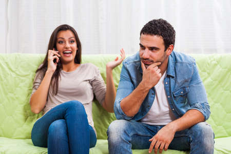 Young couple is sitting on sofa at home. Man is getting bored while woman is talking on phone.の写真素材