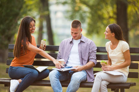 Three students are sitting on bench in park and learning.の写真素材