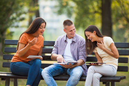 Three students are sitting on bench in park and learning.の写真素材