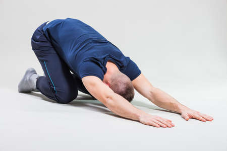 Studio shot image of young man who is stretching his body. Shashankasana / Hare pose.