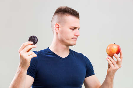 Studio shot image of young sporty man who is deciding whether to eat apple or candy.の写真素材