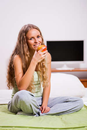 Young woman is sitting in bed and eating apple.の写真素材