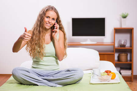 Young woman sitting in her bed and talking on phone. It's time for breakfast.の写真素材