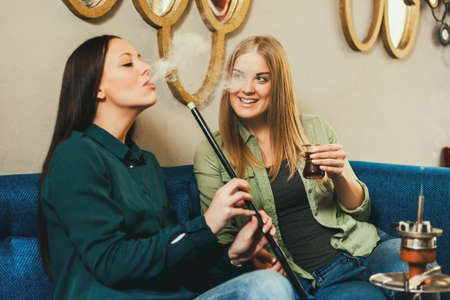 Two happy women are sitting in shisha bar and smoking nargile
